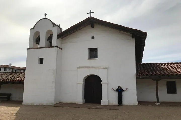 A person standing with arms outstretched in front of a white mission building.